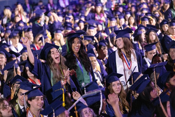 A photograph of hundreds of students cheering and smiling at graduation, wearing blue caps and gowns 