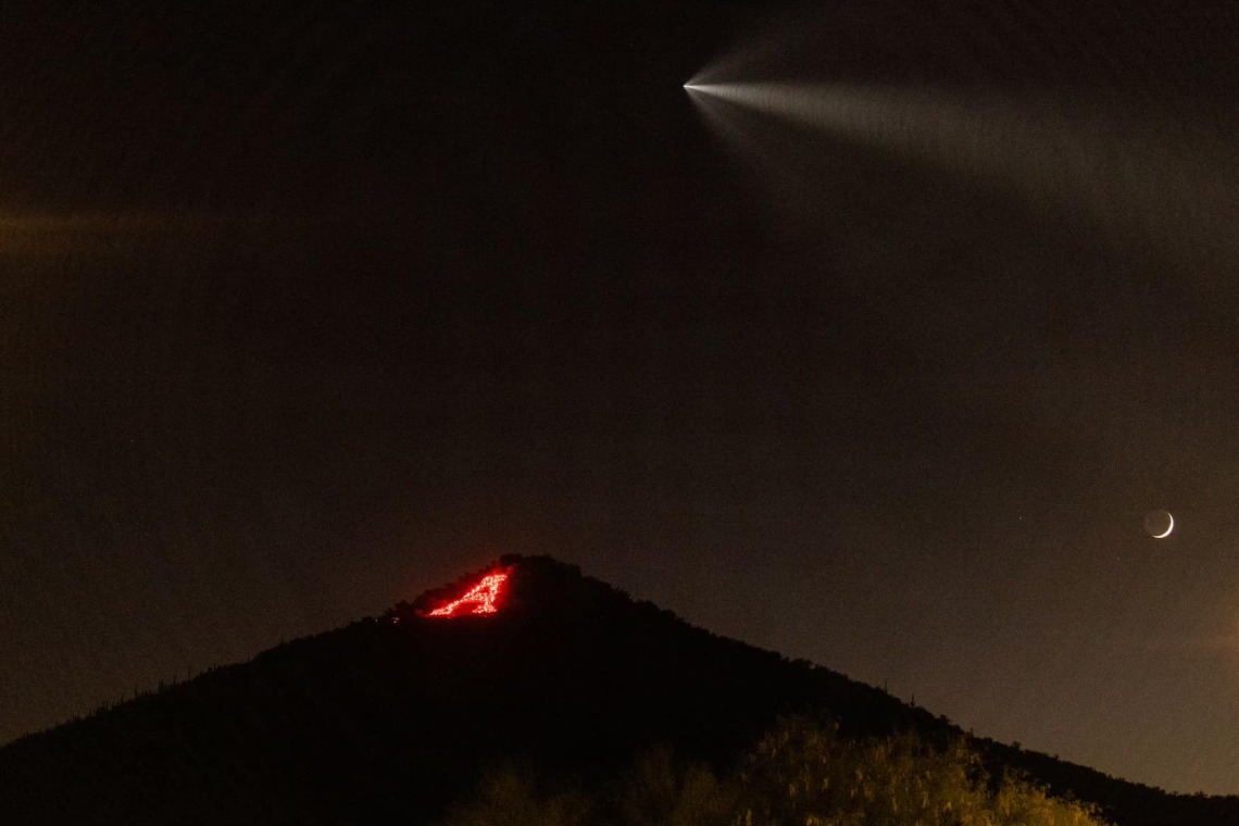 The "A" on Sentinel Peak lit up with flares as a SpaceX rocket flies above