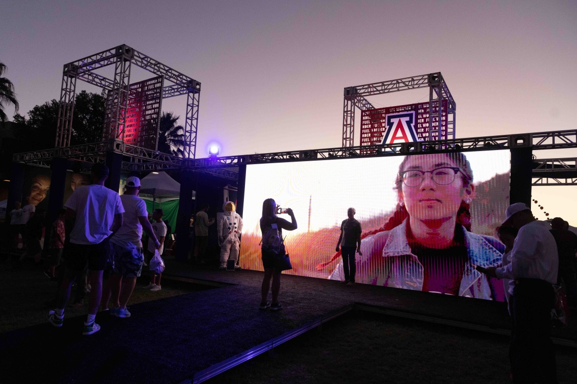 Event attendees watch large screen of video content on outdoor stage at sunset