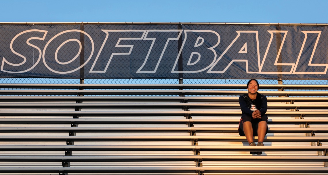 A photograph of Alyssa Palomino sitting on bleachers with the words "softball" on the banner behind her