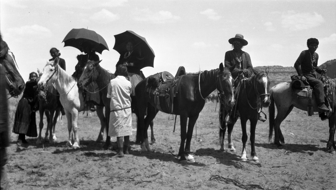 A photograph of men and women sitting either on horses or standing up. Some of the individuals are carrying umbrellas. 