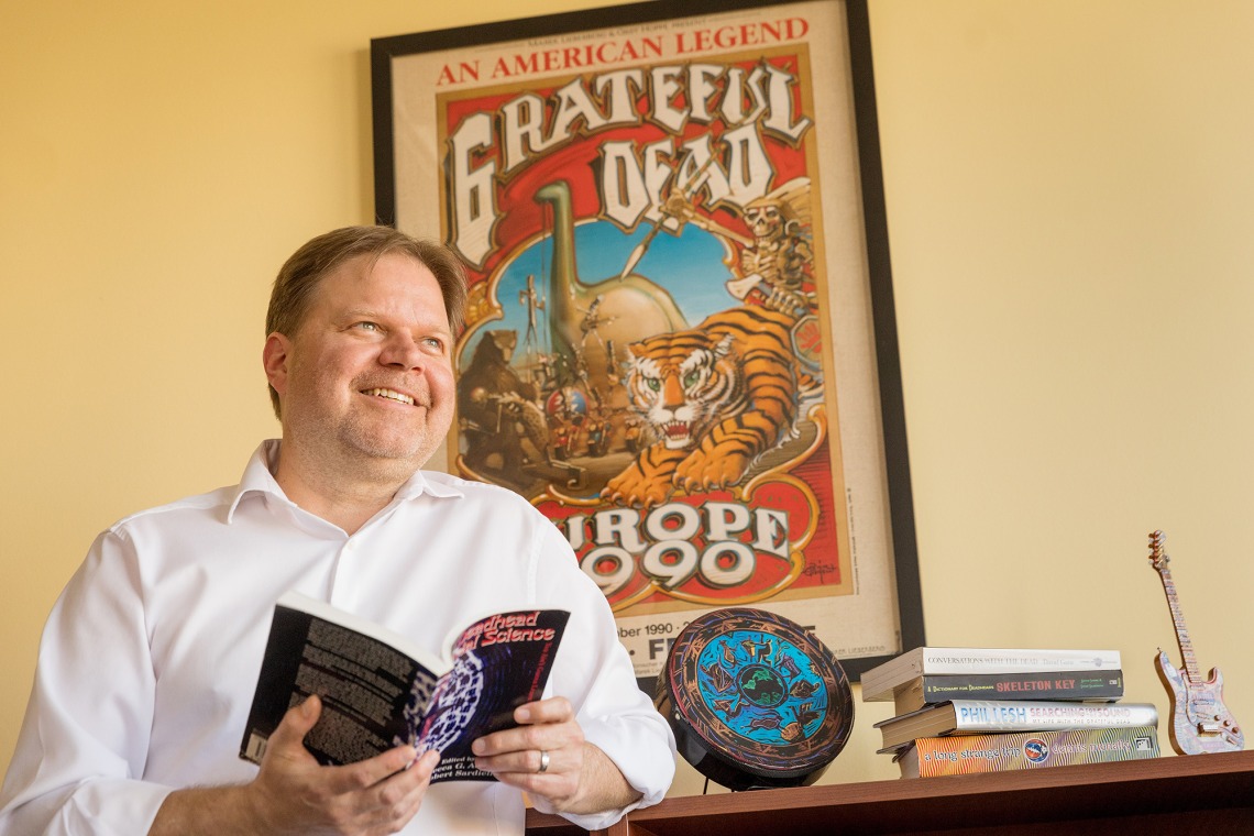 A photograph of Dean Shan Sutton, holding a book and smiling. Behind him is a "Grateful Dead" poster.