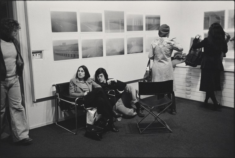 A black and white photo of individuals walking throughout a gallery installment and two others are sitting, lounging in a chair