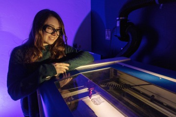 A photograph of a student, looking down and smiling, at a laser cutter carving a wildcat 